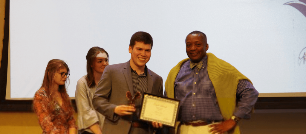 Dean Rucker stands with three students on a stage. The male student standing next to Dean Rucker is holding a framed certificate and an award.