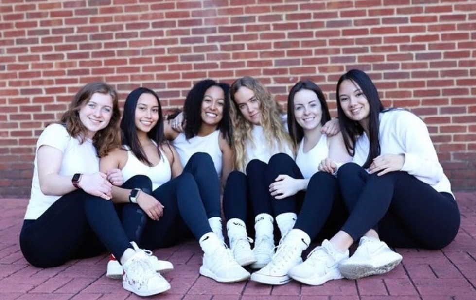 A group of girls sitting on a brick floor. They are all wearing matching white workout tops and black leggings.
