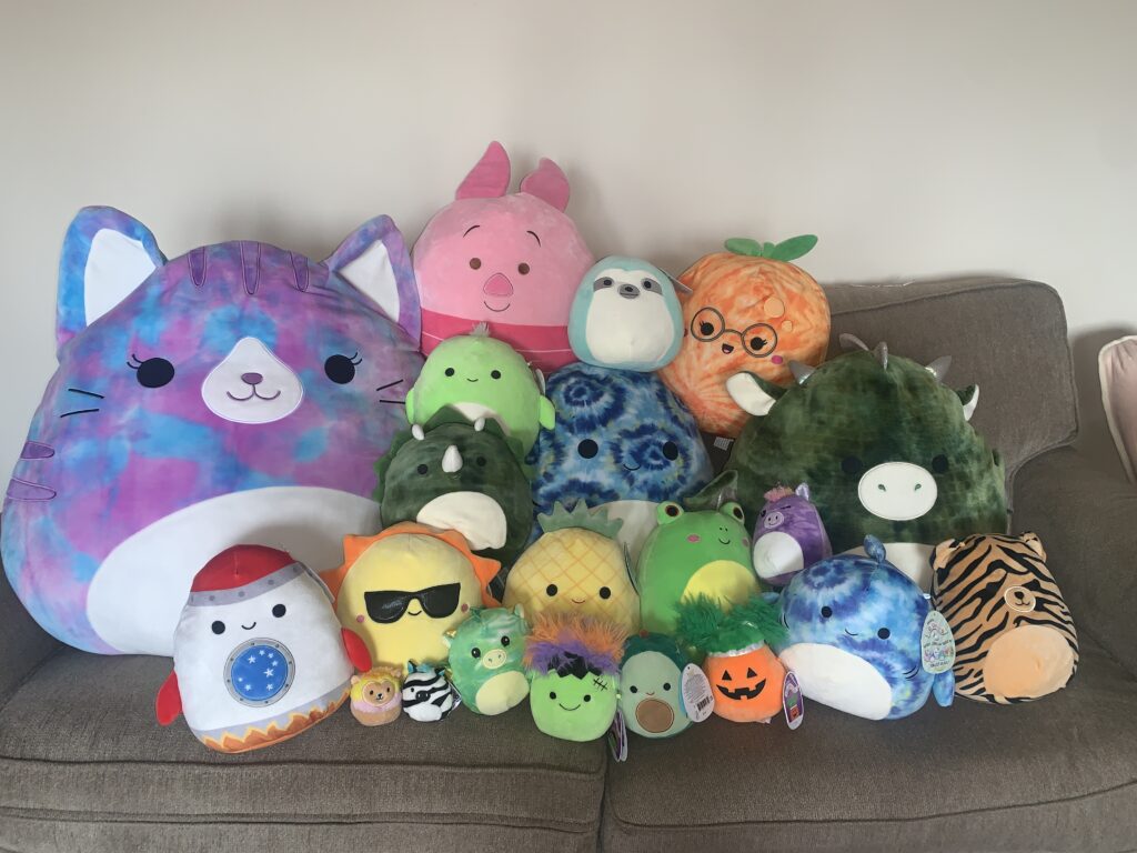 Many colorful stuffed animals lined up on a couch. They are different fruits, animals and objects with smiling faces.