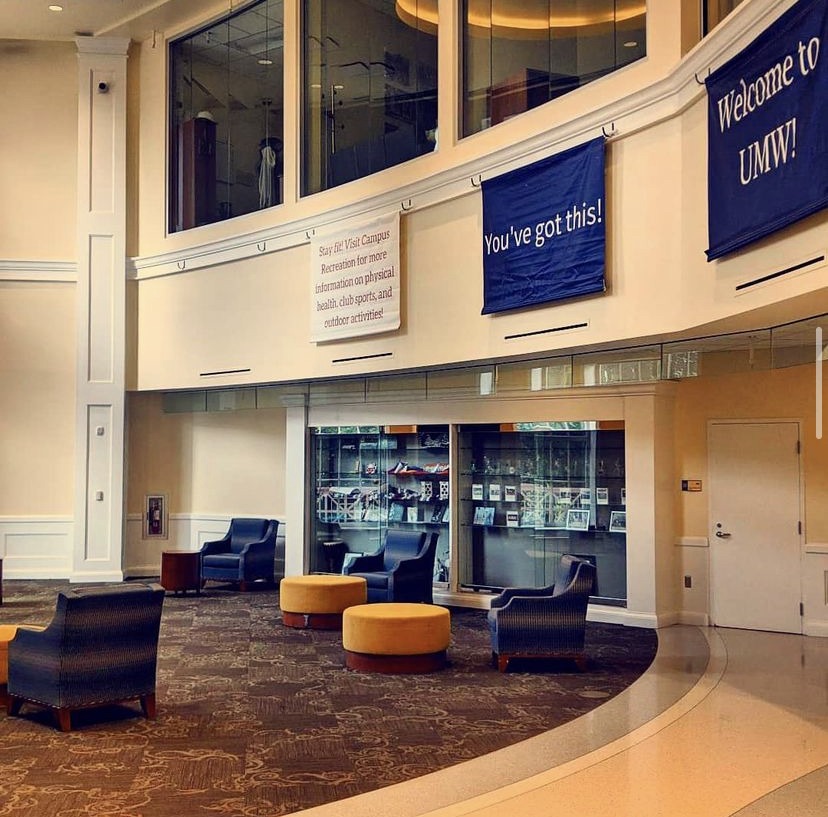 Pictured is an open room with a semi-tiled, semi-carpeted floor. On the left side are several blue armchairs and yellow ottomans. Three banners line the upper wall from left to right, which read, "Stay fit! Visit Campus Recreation for more information on physical health, club sports, and outdoor activities!","You've got this!" and "Welcome to UMW!"