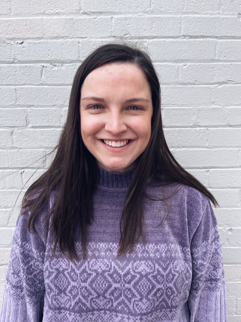 A girl with dark hair and a purple sweater is smiling at the camera.