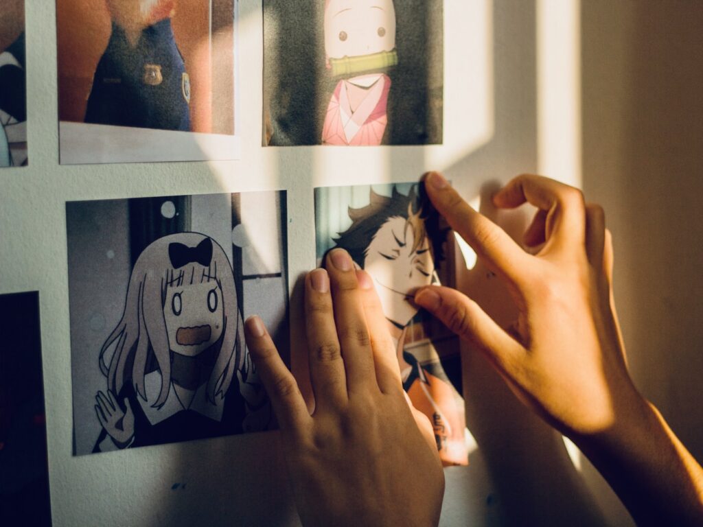 A person's hands post a picture of an animated character to the wall. The photo rests underneath an equally as small photo of a small monk, to the right of a young girl with a black bow in her hair, and diagonally down from a police officer.