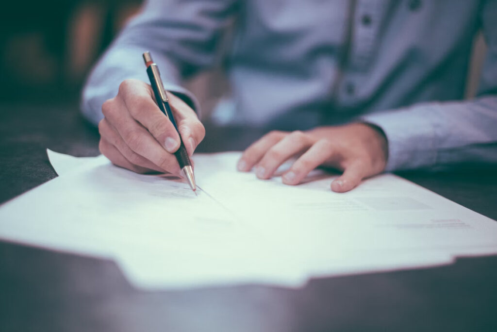 A pair of men's hands are pictured signing a small stack of papers.