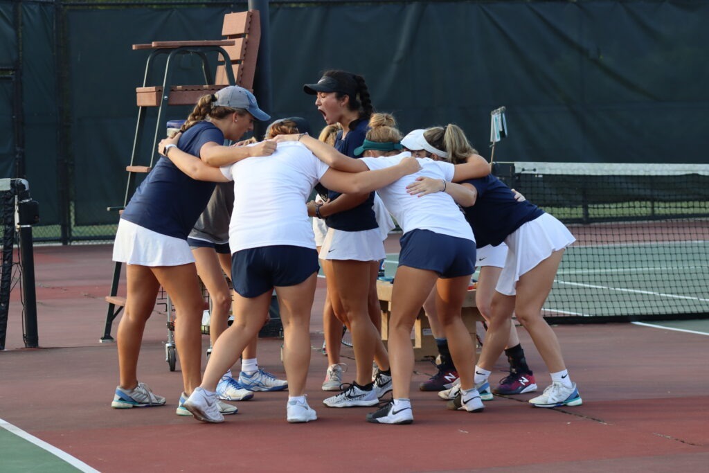 A group of female tennis players are huddled around one player in a black visor, who is yelling passionately.
