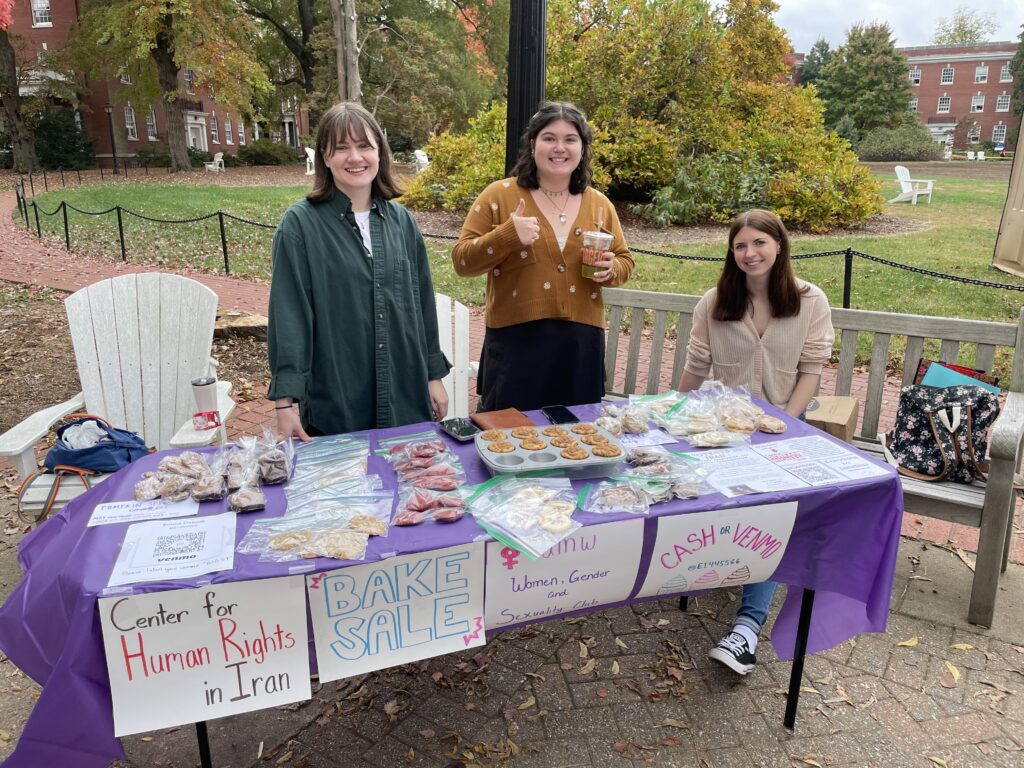 Three students smile behind a table. The table is covered in sweet treats