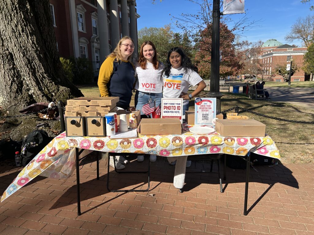 Three students pose, smiling, behind a table. The table has coffee and donuts and American flags.
