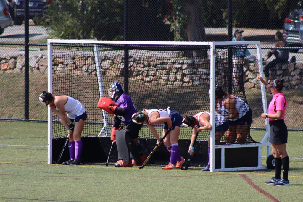 Five field hockey players stand in a field hockey goal. A referee in a pink shirt stands beside the goal. All are female presenting.
