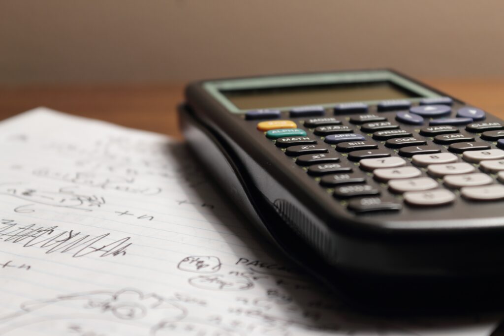 A TI-83 calculator sits on a wood surface next to a sheet of white paper containing writing.