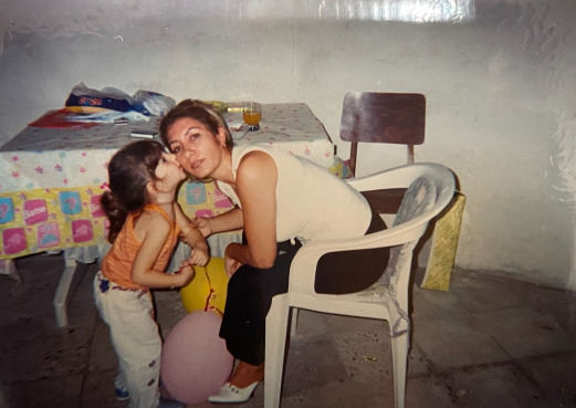 A woman sitting in a white chair at a table leans down towards her daughter to accept a kiss on the cheek.
