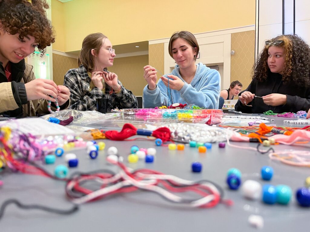 Four students sitting at a table string colorful beads on thread to make jewelry.