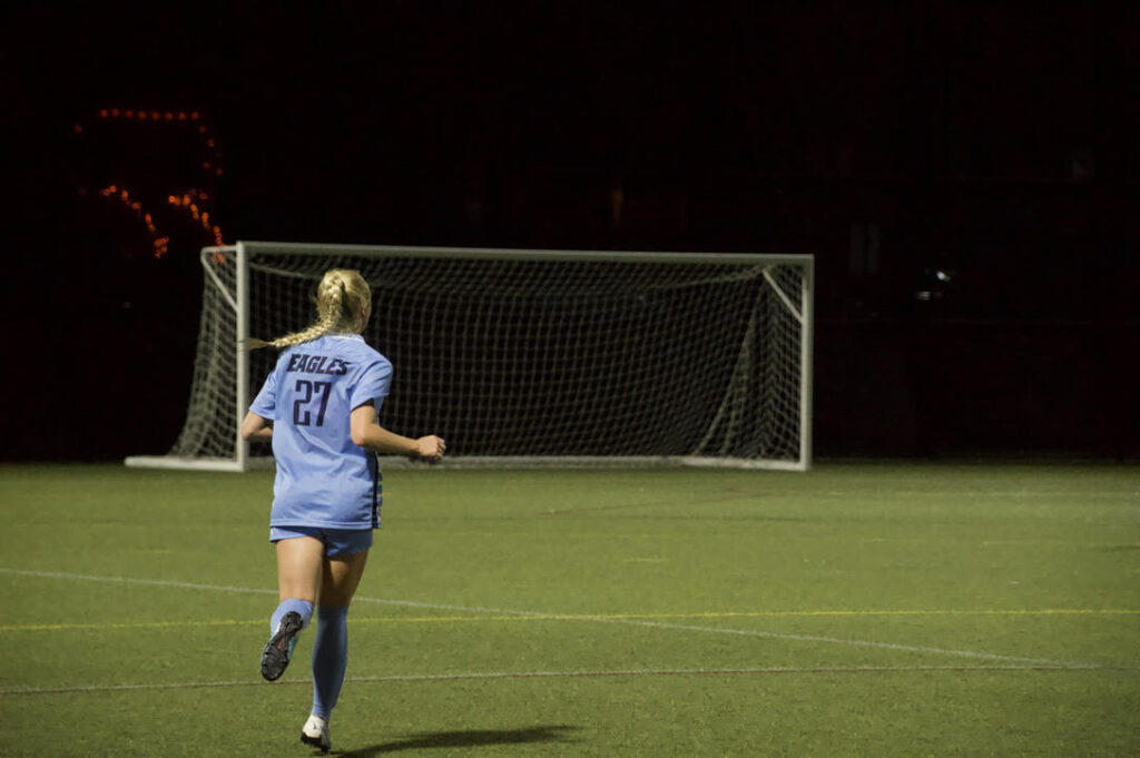 A blonde female soccer player with a light blue jersey runs toward the goal on a soccer field