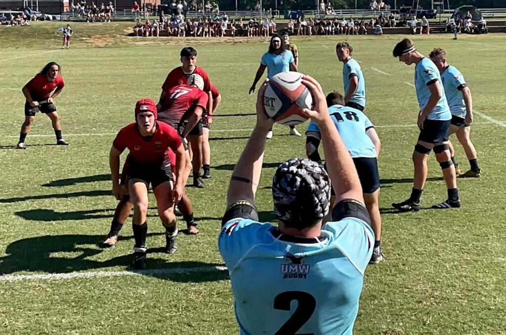 A shot of a green rugby pitch angled from behind a player carrying the ball in front of a group of teammates and opposing players.