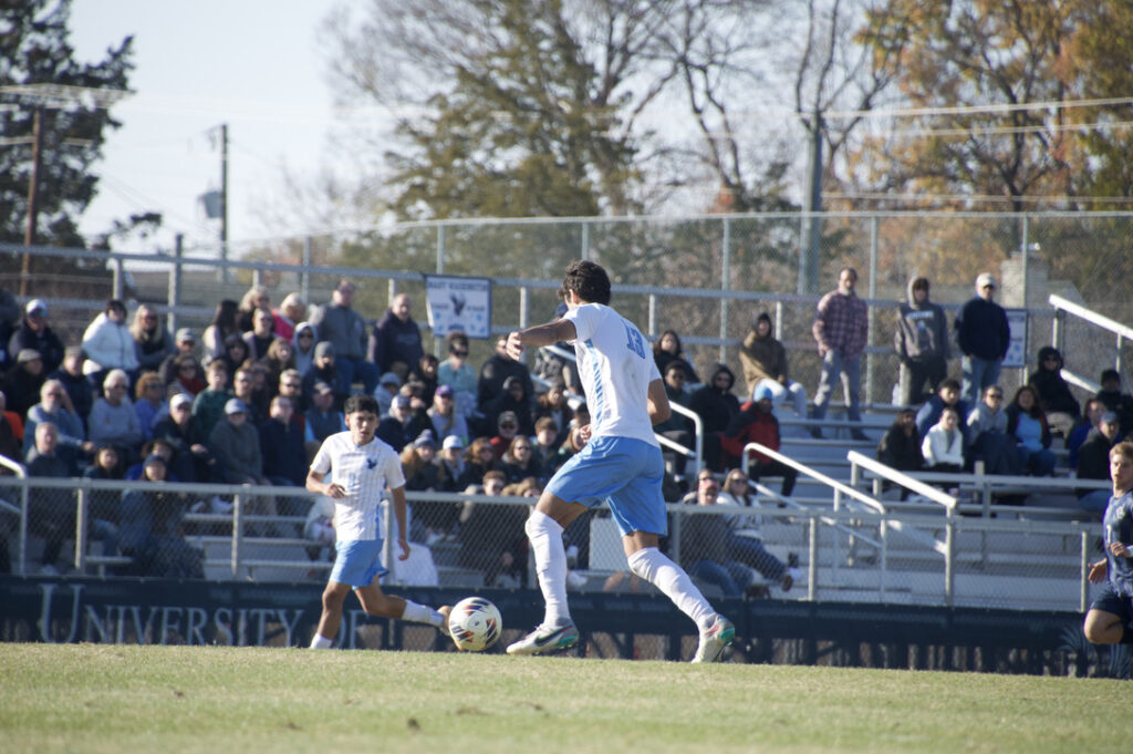 Male soccer player runs with the ball through the field as the crowd, sitting in the bleachers watches in the background.