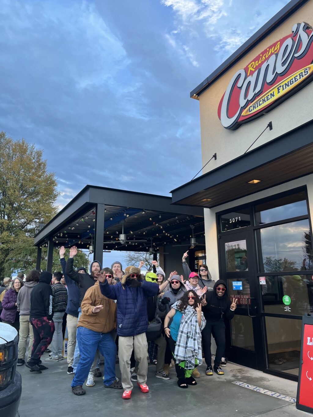 A large group of people in warm clothing outside the popular restaurant chain, Raising Cane's, celebrating and in a happy mood.