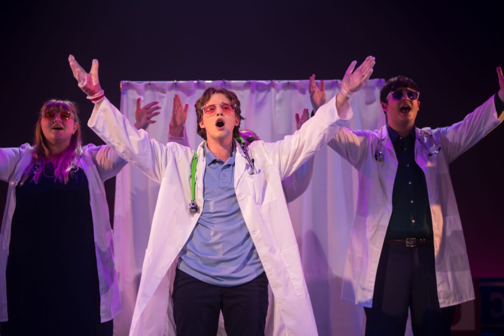 Four people dressed up as doctors singing and acting with a white curtain behind them.