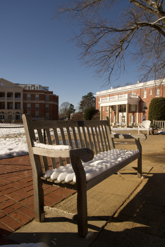 A bench covered in snow that's placed right in front of Ball circle with Virginia Hall in the background.