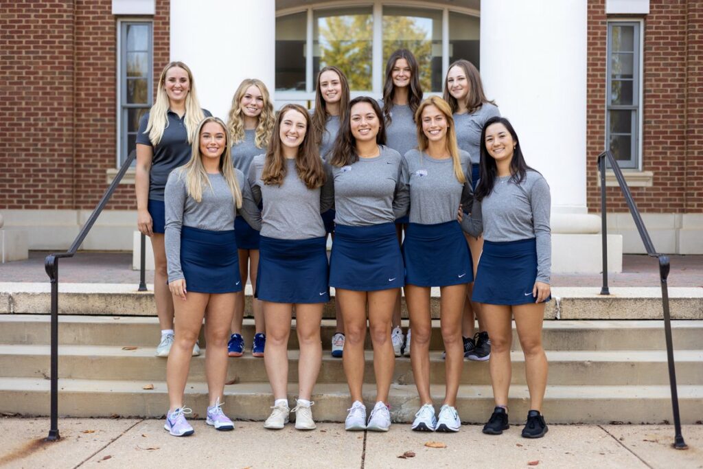 A group of women's tennis players wearing gray long sleeve tops and navy blue skirts smile and pose professionally in a group photo together in front of a brick building.