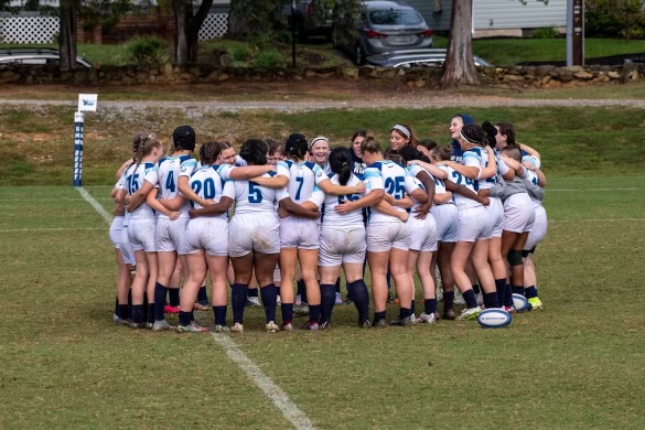 A large group of rugby girls huddling together in a scrum as they stand on the green field smiling.