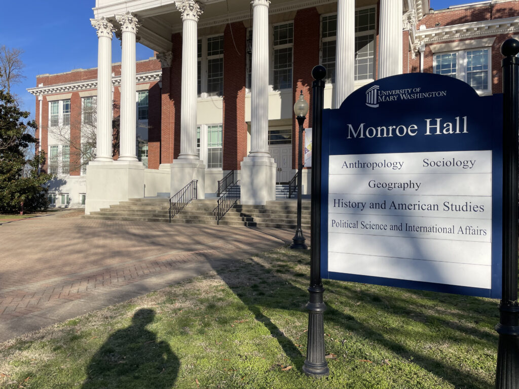 Large building with columns stands in the background of a large blue and white academic sign that reads, "Monroe Hall."