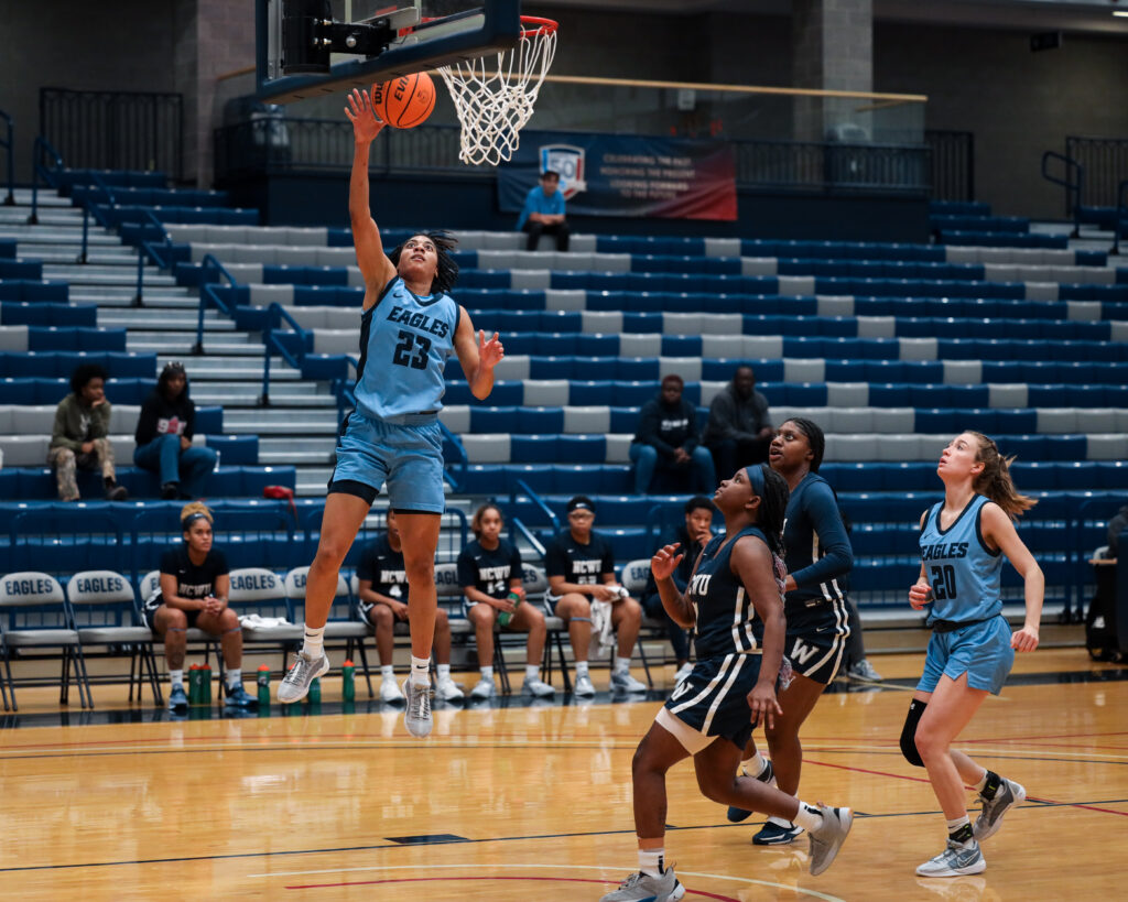 Women's basketball player jumps and scores a basket as other players race down the court behind her.