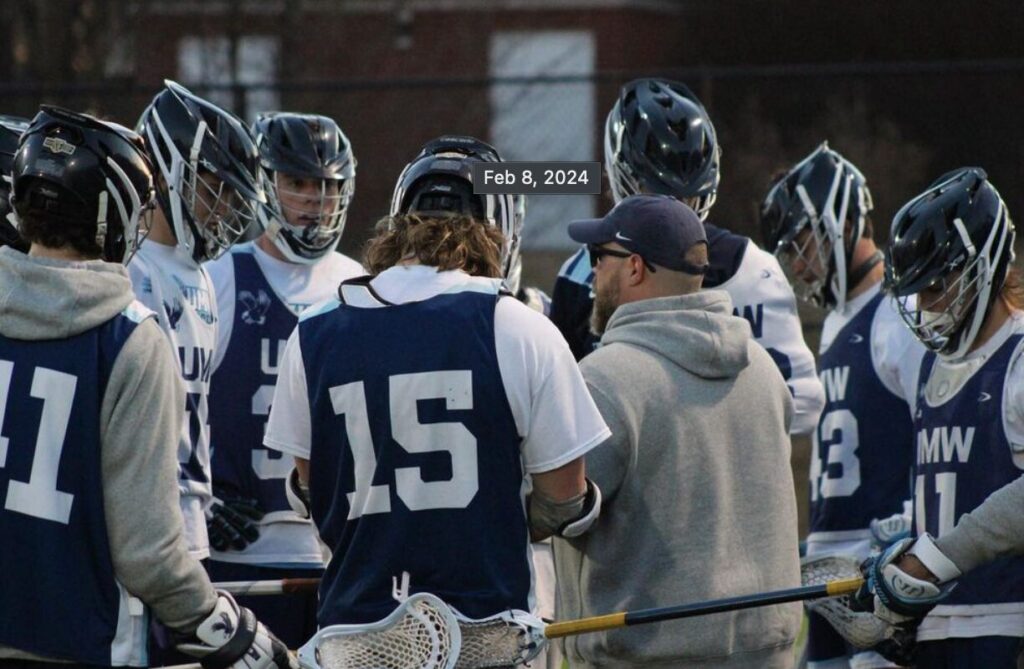 A group of lacrosse players clad in white and blue uniforms huddles with their coach on the field.