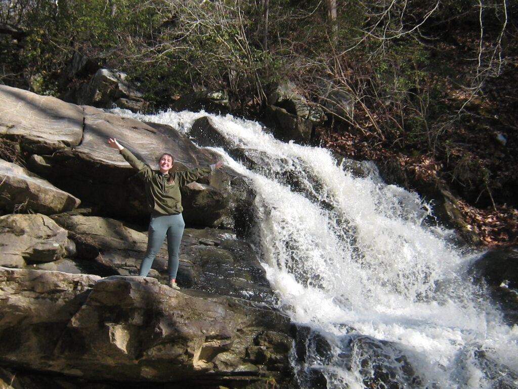 A young woman poses with her arms up next to a rushing waterfall.
