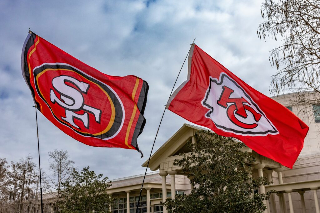 Two big red flags fly in the air next to each other. The flag on the left has the letters SF in the middle of it, representing the San Francisco 49ers football team. The flag on the right has the letters KC in the middle of it, representing the Kansas City Chiefs football team.