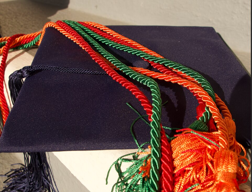 Red, orange and green graduation cords lay over a graduation cap on a step.