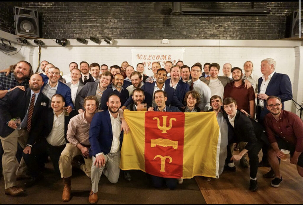 A large group of men holding up a yellow and red embroidered tapestry.