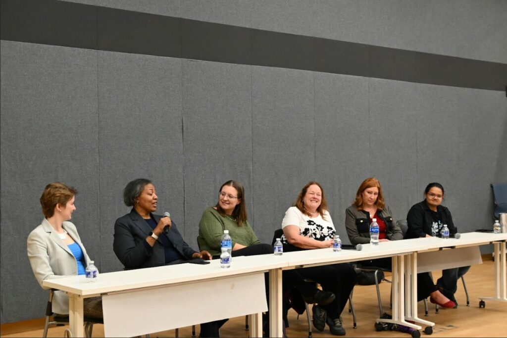 Six women speaking and looking at each other. One is speaking on a mic.