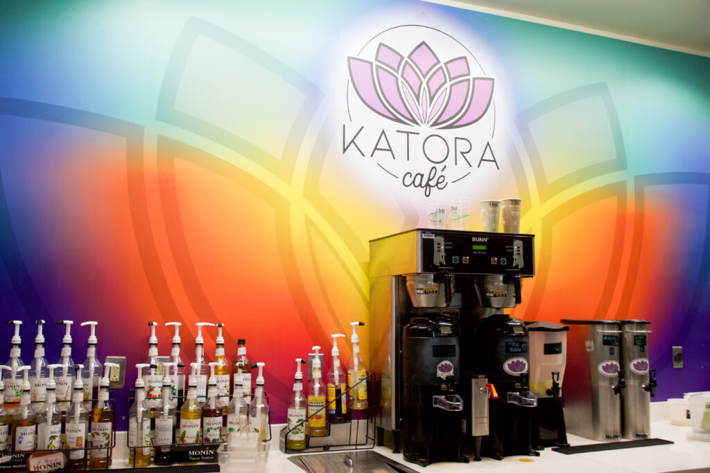 A big rainbow color Katora "sign" on the wall. The table has coffee machines and their tea packages and syrups.