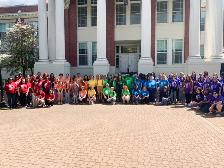 UMW community members wear red, orange, yellow, green, blue, and purple shirts in front of Monroe Hall