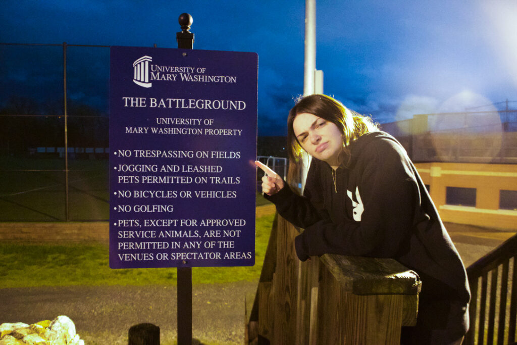 A girl looks confused as she stands and points next to a sign that says The Battleground.