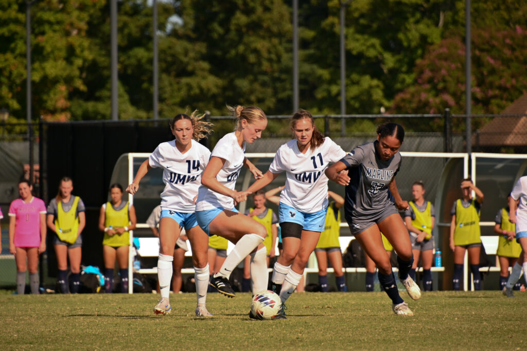 A group of three University of Mary Washington players, all facing the camera, take control of the ball as a Virginia Wesleyan University player tries to get it back from the right.