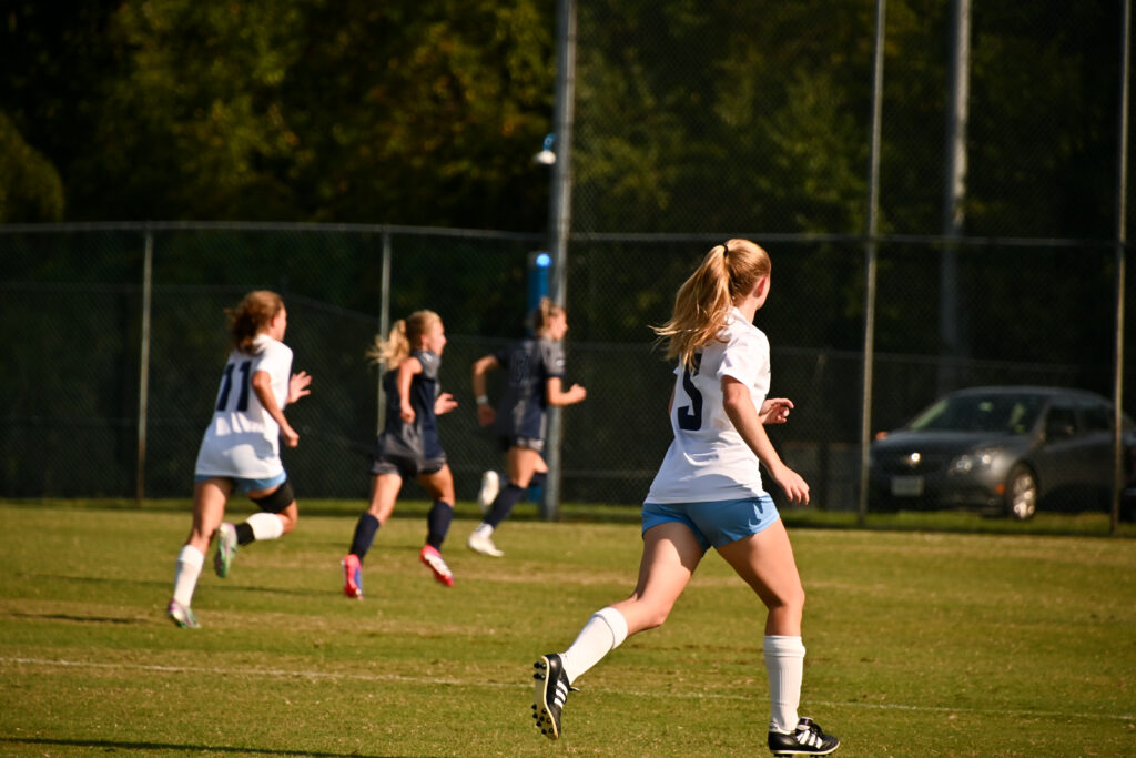 Two University of Mary Washington players, one wearing the number 5, run down the pitch with their backs to the camera as two Virginia Wesleyan University players do the same.