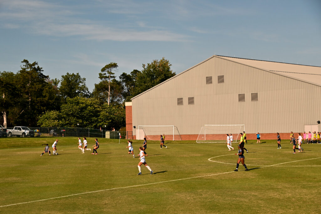 A soccer game commences between the University of Mary Washington and Virginia Wesleyan players with a large building in the background. 