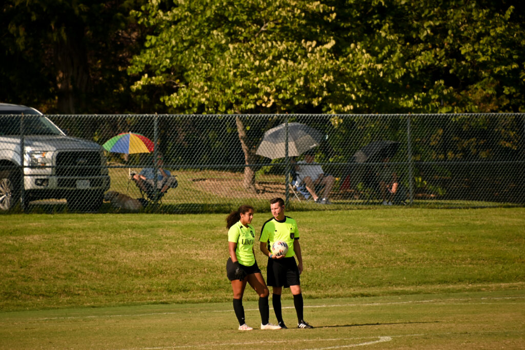 A University of Mary Washington goalkeeper stands next to a referee while they are both wearing a neon green top, black shorts and black socks.