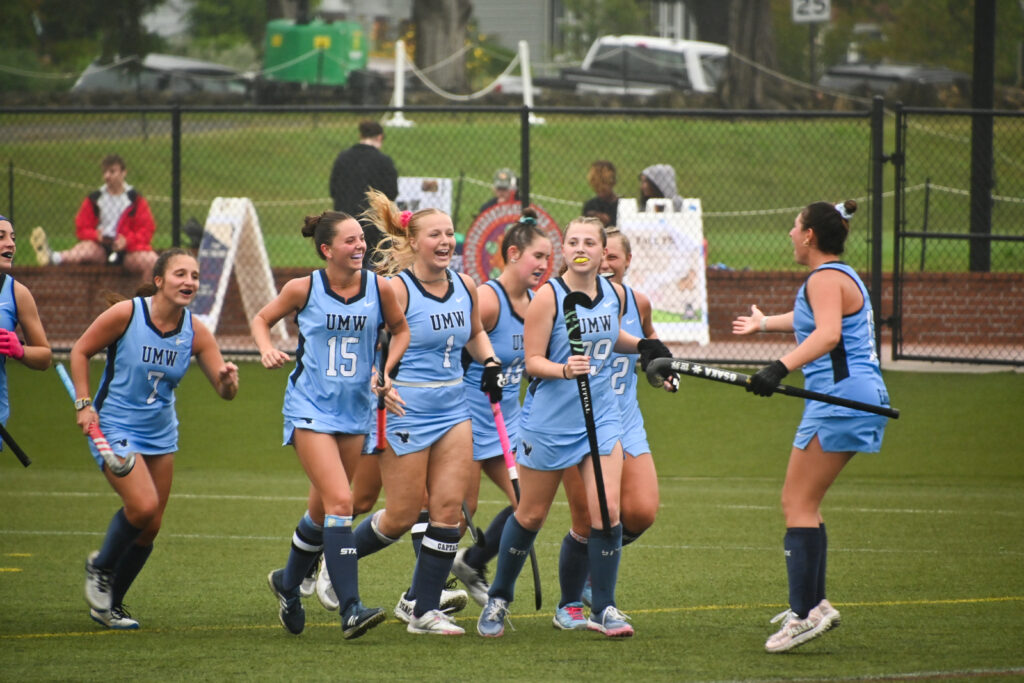 A group of Mary Washington players, all with smiles on their faces, walk towards a girl with her back to the right as she reaches out her hand for a high-five.