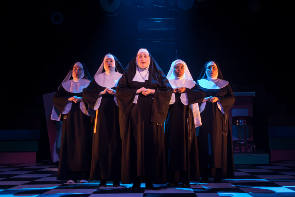 Actors dressed as nuns clasp hands together on stage.