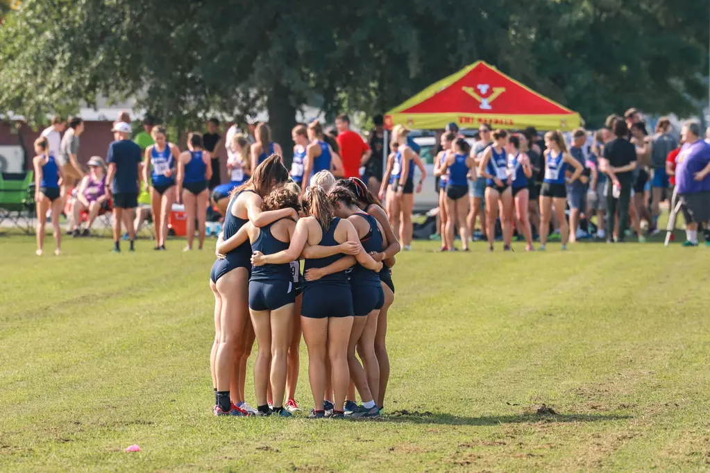 The UMW women's cross country team huddles up with their back to the camera in front of another team. They are wearing blue uniforms.